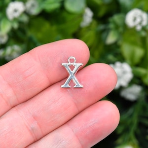 May include: Silver charm in the shape of the letter X. The charm is being held in a hand.