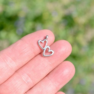 May include: A silver charm with two interlocking heart shapes. The charm is held in a hand against a green background.