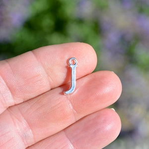 May include: Silver charm in the shape of the letter J. The charm is being held in a hand.
