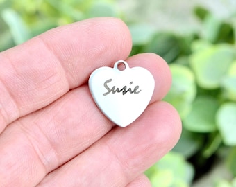 May include: A small, heart-shaped silver-toned metal charm featuring the name Susie engraved in a cursive script. The charm includes a small circular loop at the top for attachment, presenting a smooth, polished surface with dark, clear lettering.