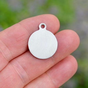 May include: A small, round, silver-colored metal pendant with a loop at the top. The pendant is held in a person's hand, and the background is blurred green.
