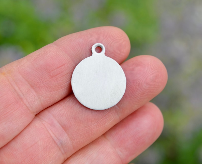 May include: A small, round, silver-colored metal pendant with a loop at the top. The pendant is held in a person's hand, and the background is blurred green.