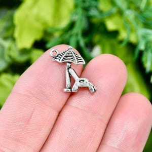 May include: Silver charm of a beach chair with an umbrella. The chair is facing to the right and the umbrella is open. The charm has a loop at the top for attaching to a bracelet or necklace.