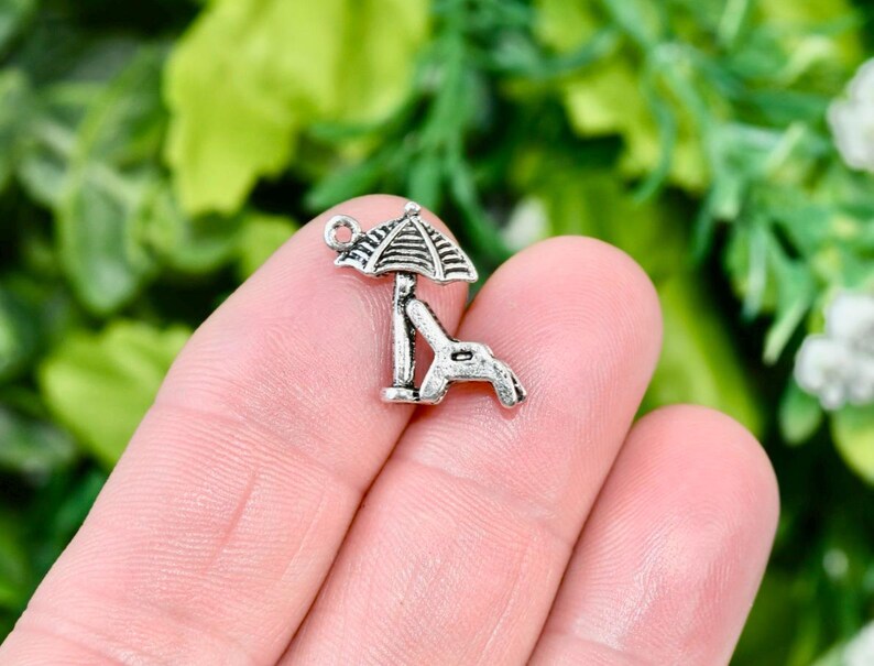 May include: Silver charm of a beach chair with an umbrella. The chair is facing to the right and the umbrella is open. The charm has a loop at the top for attaching to a bracelet or necklace.