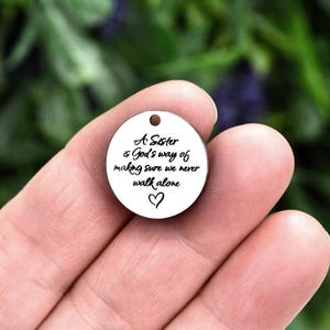 May include: A circular silver-toned metal charm featuring a small hole at the top and black engraved text that reads: A Sister is God's way of making sure we never walk alone, accompanied by a small hand-drawn heart symbol at the bottom.
