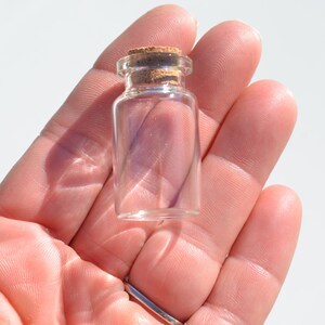 May include: A small, clear glass bottle with a cork stopper. The bottle is held in a hand, showcasing its size and design. The cork is a light brown color, contrasting with the transparent glass. Ideal for small crafts or decorative purposes.