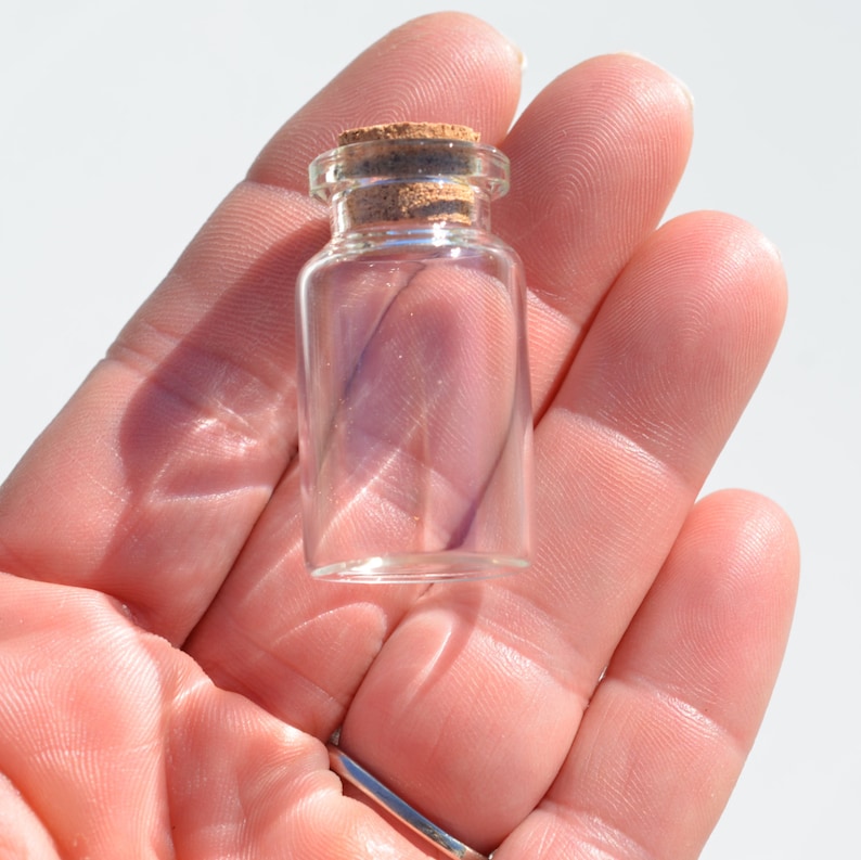 May include: A small, clear glass bottle with a cork stopper. The bottle is held in a hand, showcasing its size and design. The cork is a light brown color, contrasting with the transparent glass. Ideal for small crafts or decorative purposes.