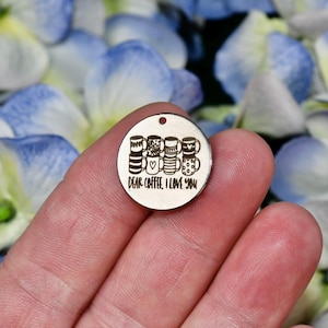 May include: A round, wooden charm with a small hole at the top. The charm features an engraved design of four coffee mugs and the text "DEAR COFFEE, I LOVE YOU". The charm is held in a person's hand against a background of blue and white flower petals.