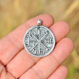 May include: Silver pendant with a circular design featuring a compass rose with runes. The pendant is held in a hand.