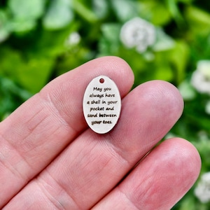 May include: A small, oval-shaped wooden tag with the text "May you always have a shell in your pocket and sand between your toes." The tag is being held in a hand.