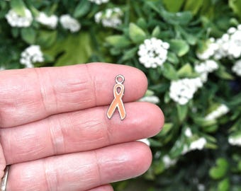 May include: A small, orange ribbon charm with a silver-colored border and a loop at the top. The charm is held in a person's hand, with a blurred background of green foliage and white flowers.