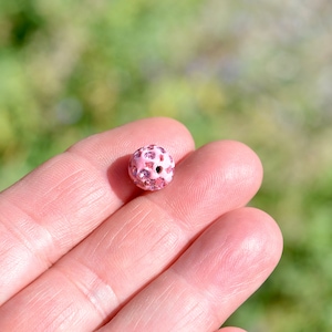 May include: A pink round bead with small pink rhinestones covering the surface. The bead has a hole in the center.