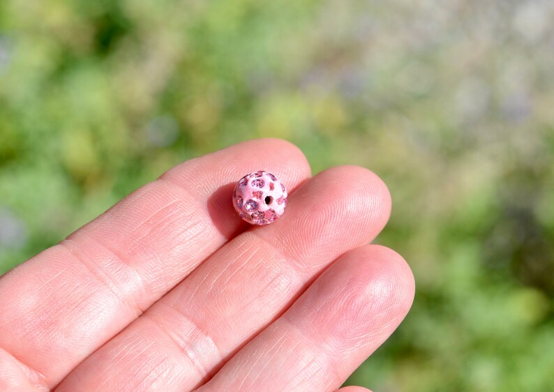 May include: A pink round bead with small pink rhinestones covering the surface. The bead has a hole in the center.