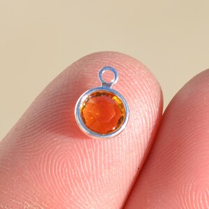 Pu&ograve; includere: Un piccolo ciondolo d'argento con una pietra preziosa rotonda di colore arancione al centro. Il ciondolo ha un anello nella parte superiore per attaccarlo a una collana o un braccialetto.