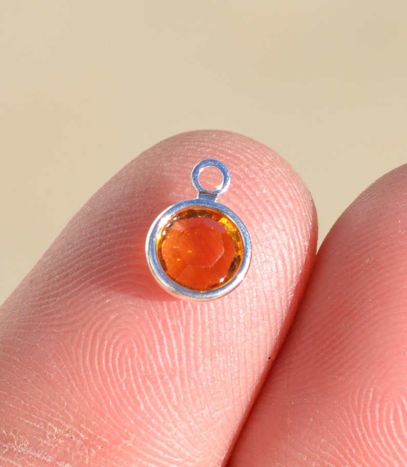 Pu&ograve; includere: Un piccolo ciondolo d'argento con una pietra preziosa rotonda di colore arancione al centro. Il ciondolo ha un anello nella parte superiore per attaccarlo a una collana o un braccialetto.