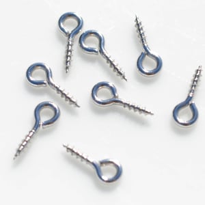 May include: Silver screw-in eyelet hooks, a type of jewellery finding, are scattered on a white surface. These small metal hooks are used for attaching jewellery components.