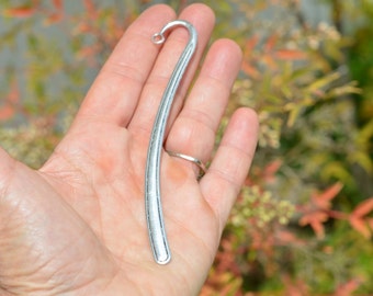 May include: A silver bookmark with a curved design. The bookmark is held in a hand.