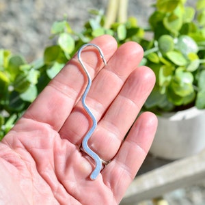 May include: A silver-colored, wavy bookmark with a hooked top and a rounded bottom. The bookmark is held in a hand, with a blurred background of green foliage and a white pot.
