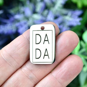 May include: A rectangular, off-white charm with the word "DADA" printed twice in black, within a black border. The charm has a small hole at the top for attachment. The background is blurred with green and purple foliage.