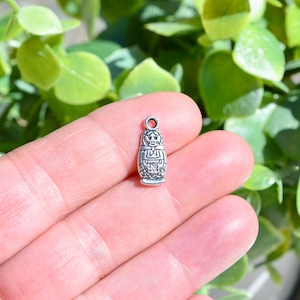 May include: Silver charm in the shape of a traditional Russian nesting doll. The charm has intricate details and a loop for attaching to a necklace or bracelet.