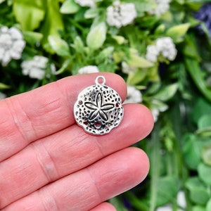 May include: Silver charm in the shape of a sand dollar with a five-pointed star in the centre. The charm has a textured surface and is ideal for jewellery making.