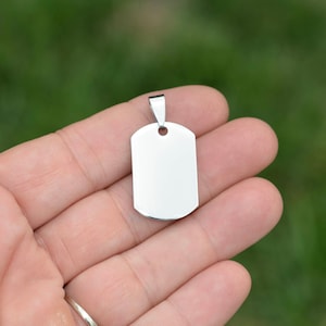 May include: A silver blank dog tag pendant with a rounded rectangular shape and a small loop at the top.