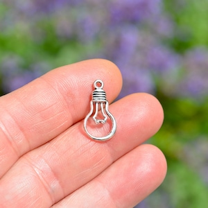 May include: Silver light bulb charm pendant. The charm is in the shape of a traditional light bulb with a filament inside. The charm is on a plain background.