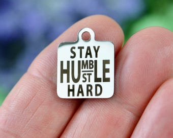May include: A square silver-toned metal charm with rounded corners and a small circular loop at the top. The surface features black engraved text that reads STAY HUMBLE HUSTLE HARD, with the word HUMBLE stacked vertically in the center.