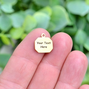 May include: Gold-coloured metal charm with a rounded rectangular shape. The charm has a hole at the top for attaching to a necklace or bracelet. The charm is engraved with the text "Your Text Here".