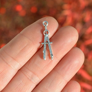 May include: Silver compass charm with a detailed design. The compass is open and the points are sharp.