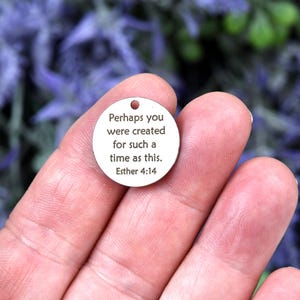 May include: A small, round, silver-colored charm with a hole at the top. The charm has the quote "Perhaps you were created for such a time as this. Esther 4:14" in black text. The charm is held in a person's hand, with a blurred background of purple flowers.