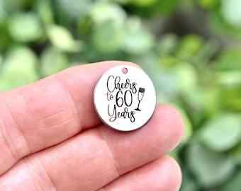 May include: A round, silver-colored charm with the text "Cheers to 60 Years" in black script. A champagne flute graphic is also included. The charm has a small hole at the top for attaching to a necklace or bracelet.