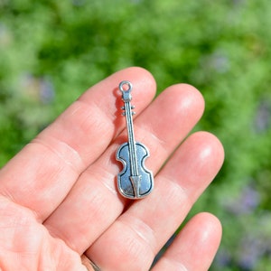 May include: A silver-colored violin charm with a loop at the top for attaching to a necklace or bracelet. The charm has a detailed design, including the strings and the shape of the instrument. The background is a blurred green and purple.