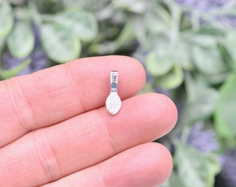 May include: A small, silver-colored pendant with a textured, oval-shaped base and a rectangular loop at the top. The pendant is held between fingers against a blurred background of green and purple foliage.