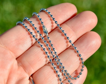 May include: A silver ball chain necklace with a lobster clasp closure. The chain is made of small, round beads.