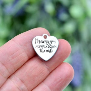May include: A silver-toned heart-shaped metal charm featuring a small circular hole at the top for attachment. The surface is engraved with the black text "Missing you AS I WALK DOWN the aisle" in a combination of script and sans-serif fonts.
