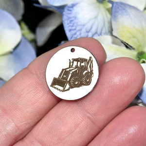 May include: A circular silver-toned metal charm featuring a dark, laser-engraved illustration of a construction tractor with a front loader and backhoe. The charm has a small hole at the top for attachment, displaying a detailed heavy machinery design.