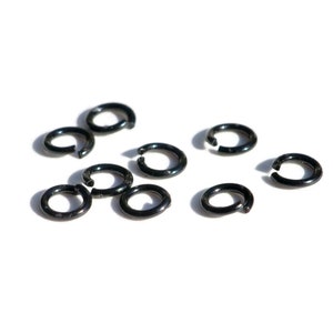 May include: A group of ten black metal jump rings, scattered on a white background. The rings are all the same size and shape, and they are perfect for jewelry making or other crafts.