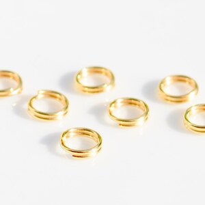 May include: Eight gold jump rings, also known as split rings, are arranged in a semi-circle pattern on a white surface. These rings are commonly used in jewellery making.