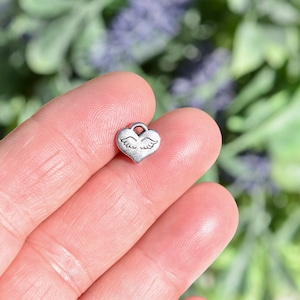 May include: A silver heart-shaped charm with a pair of wings etched into the surface.