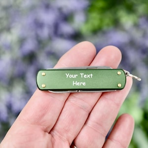 May include: A green pocket knife with a silver blade and a gold-colored ring. The knife has the text "Your Text Here" engraved on the handle.