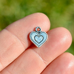May include: Silver charm in the shape of a heart with a smaller heart inside. The heart has a dotted border.