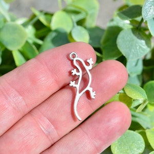 May include: Silver-toned metal charm shaped like a lizard. The charm is detailed with a cutout design and has a loop at the top for attaching to a necklace or bracelet.