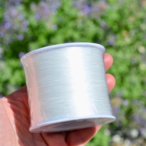 May include: A spool of clear elastic thread, wound around a white plastic core. The thread is translucent and appears to be very thin.