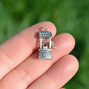 May include: Silver charm in the shape of a well with a bucket and a handle. The charm is detailed with a roof and a textured base.
