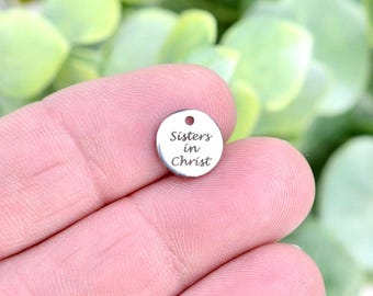May include: A small, circular, silver-toned metal charm featuring a polished finish and a single hole at the top. The text "Sisters in Christ" is engraved in a dark, cursive font on the front surface of the round metal disc.