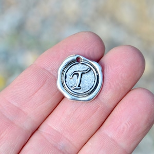 May include: Silver charm with a wax seal design featuring the letter "T" in a cursive font.