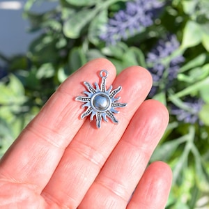 May include: Silver charm in the shape of a sun with a textured surface. The sun has a round centre and rays extending outwards.