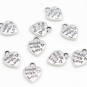 May include: A collection of small, silver-colored heart charms. Each charm has the words "made with love" engraved on it. The charms have a loop at the top for attaching to jewelry or other crafts. The charms are scattered on a white surface.