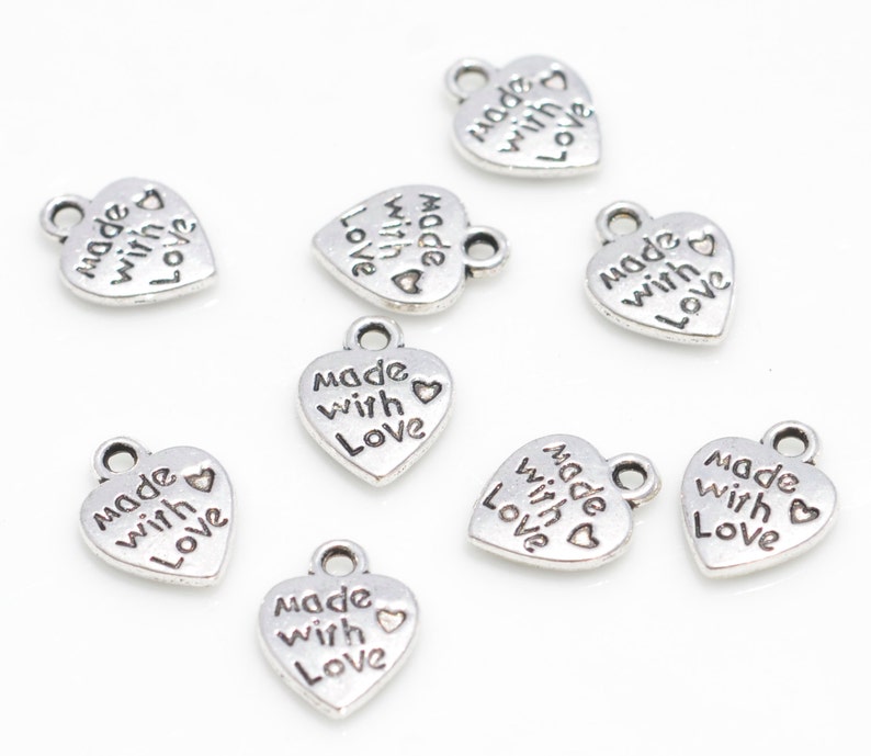 May include: A collection of small, silver-colored heart charms. Each charm has the words "made with love" engraved on it. The charms have a loop at the top for attaching to jewelry or other crafts. The charms are scattered on a white surface.